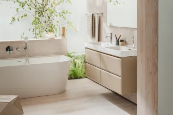 Bright modern bathroom with a freestanding white tub, wood-toned double vanity, large mirror, towels, and lush indoor plants beside a tree against a wooden wall.