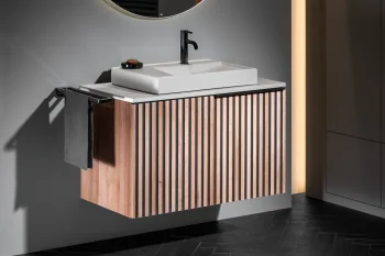 Modern bathroom with a white rectangular sink on a wooden slatted vanity, black faucet, circular backlit mirror, grey walls, dark towel rail and warm vertical lighting.