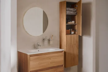 Round mirror above a white ceramic sink set into a light wood vanity, with an open wall cabinet displaying towels and toiletries.