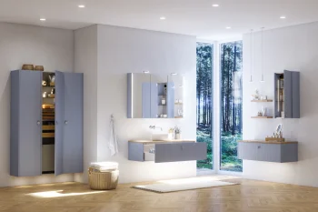 Modern bathroom with blue-grey cabinets and floating vanities, mirrored storage, a towel, wicker basket, and a large forest-facing window over a wood floor.