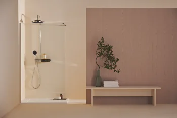 Minimalist bathroom with a glass shower and beige walls, plus a light wood panel backdrop. A bench holds a green vase with foliage and white towels.