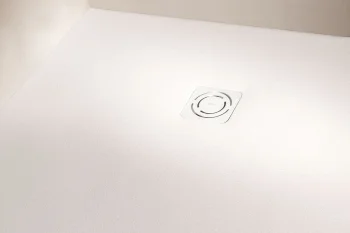 Corner of a pale white ceiling with a square white vent cover featuring a circular grille, seen at an angle where two walls meet.