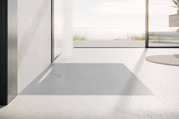 Minimal bright bathroom with a large rectangular grey shower tray on pale floor, glass wall to a sunny deck with a sea view and a round rug nearby