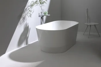 Minimalist white bathroom with a freestanding oval tub, chrome tap, glass shelf, and a tall plant casting shadows on a bright angled wall, plus a white chair.