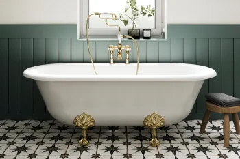 White freestanding bathtub with ornate gold feet and brass taps, set against green wainscoting, a patterned tiled floor, a window, plants, and a flamingo print.