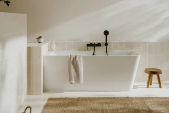 Minimalist bathroom with a white freestanding tub under a sloped ceiling, beige tiles, black fixtures, a towel over the tub, a plant on the left, a wooden stool and woven rug.