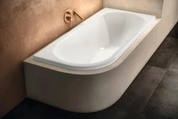 White, modern bathtub built into a beige surround with a copper wall-mounted faucet on a warm brown wall, and a dark stone floor.