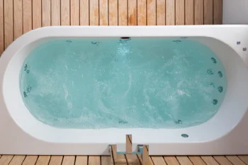 White freestanding bathtub filled with bubbly turquoise water, set on a wooden slatted floor and walls, with a plant visible in the corner.