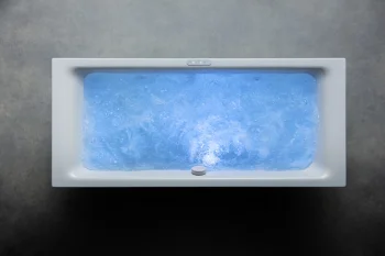 Top-down view of a rectangular white bathtub filled with swirling blue water, with a drain and controls along the edge, on a dark grey floor.