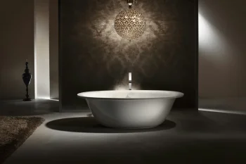 White freestanding bathtub in a dim bathroom with a patterned wall, a spherical chandelier overhead, tall black vase to the left, and a shaggy rug.