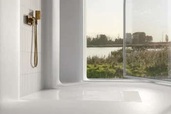Bright minimalist bathroom with a gold handheld shower mounted on white tiles, beside a large open window framing a river and trees outside.
