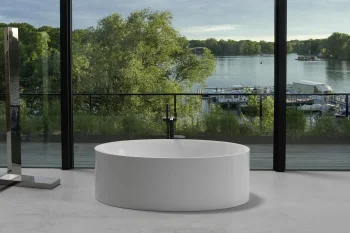 White cylindrical freestanding bathtub in a bright modern bathroom with floor-to-ceiling glass, overlooking a lake with trees and moored boats.