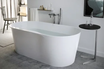 Modern bathroom with a white freestanding tub half-filled with water, chrome tap, a black round side table with glass decanters, and grey walls.