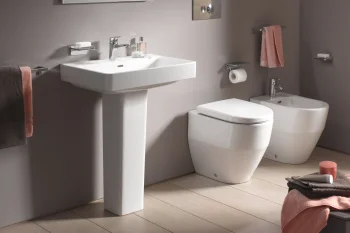 Modern bathroom with a white pedestal sink, backlit mirror, toilet and bidet, taupe walls, pink towels on rails, and a light-wood floor.