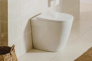 Minimalist bathroom with a white floor-standing toilet against pale tiled wall; a flush plate, toilet paper on a holder, a woven basket and a beige rug nearby.