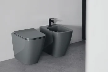Modern bathroom with a grey toilet and matching grey bidet on a concrete floor; black faucet on the bidet and a black wall flush panel.