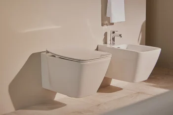 Bathroom with a wall-mounted white toilet and a bidet, chrome taps, a white towel on a wall rack, beige walls and a light tiled floor.