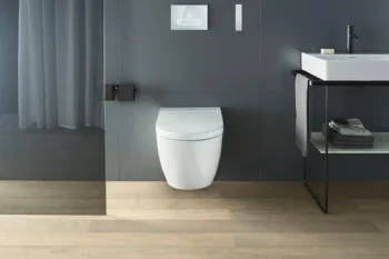 Modern bathroom with a wall-mounted white toilet, grey panels, a white sink on a metal frame, pale wooden floor, and a sheer curtain on the left.