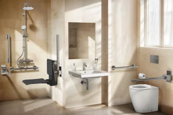 Light-filled beige bathroom featuring a wall-mounted sink with mirror and soap dispenser, accessible shower with fold-down bench and grab bars, and a toilet with rails.