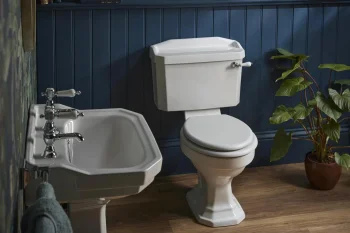 A bathroom with dark blue beadboard walls and tropical wallpaper, a white toilet and pedestal sink, wooden flooring, and a potted plant by a shelf of decor.