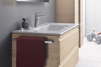 Modern bathroom with a floating oak vanity, white sink and chrome faucet, backlit mirror, tall storage cabinet, burgundy towel, grey walls and tiled floor.