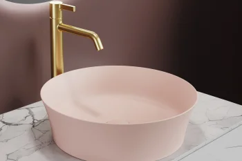 A soft pink circular sink on a white marble counter with a tall gold faucet, set against a pink gradient wall.