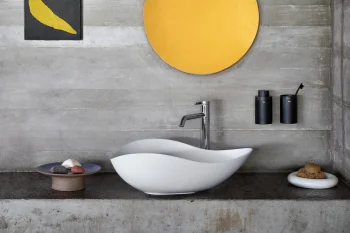 Minimalist bathroom with a curved white sink and chrome tap on a concrete wall; a large yellow circular mirror, a small dark square artwork with a yellow shape, and black wall-mounted dispensers.