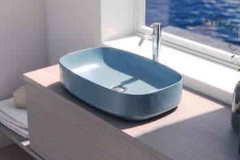 Light blue oval sink on a pale wooden vanity by a window overlooking blue water; chrome faucet, folded towels and a plant on the counter.