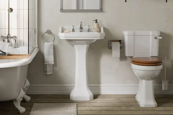 White pedestal sink, clawfoot tub to the left, toilet with wooden seat, towel ring, and a bath tray with a book on a neutral wood floor.