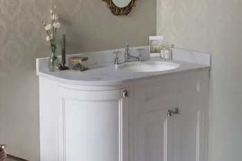 White bathroom vanity with marble top under an ornate gold-framed oval mirror; damask wallpaper, glass-and-gold side table, and decorative items.