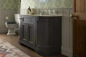 Dark grey bathroom vanity with white top and chrome faucet against green damask wallpaper; toilet to the left and wooden floor with a door to right.