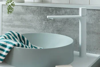 Modern bathroom sink with round pale-blue basin, white tall faucet, teal-striped towel, soap dish, and a leafy plant against white paneled wall.