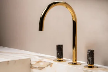 Shiny gold curved faucet with two black marble-topped handles on a white marble sink, set against a beige wall with a partial white-framed mirror above.