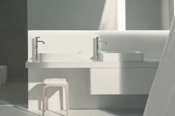 Minimalist white bathroom with two rectangular vessel sinks and chrome taps on a floating counter, a stool with a folded towel, and a curtain on the right.