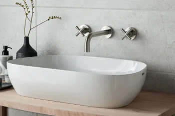 White oval basin on a wooden counter with a chrome wall‑mounted faucet, round mirror above, black vase with stems, and a soap dispenser.