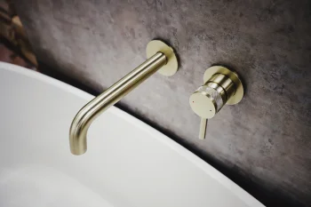 Brass wall-mounted bath tap with curved spout and matching knob above a white tub, set against a textured grey wall.