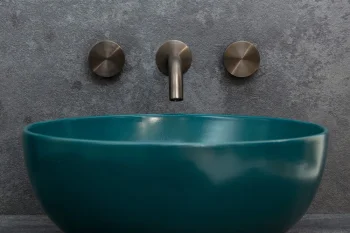 Teal round ceramic sink basin beneath a brushed metal wall-mounted faucet with three circular handles on a grey textured wall.