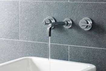 Chrome wall-mounted faucet with two round handles on a blue-grey tiled wall, water flowing into a white rectangular sink; grooming items at the edge.