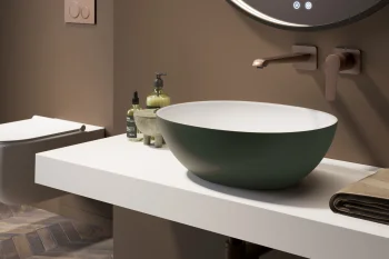 Modern bathroom with a round backlit mirror, white floating counter, green bowl sink, wall-mounted tap, soap bottles, towel, and herringbone floor.