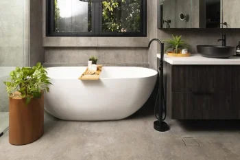 Modern bathroom with a white freestanding bathtub, wooden bath tray, potted plants, black floor-mounted tap, grey concrete walls, and a vanity with dark cabinets.