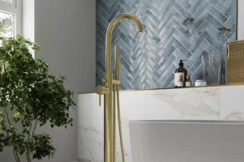Brass freestanding bathtub tap with a handheld hose beside a white tub, blue chevron tiled wall, marble surround, and a potted plant in the bright bathroom.
