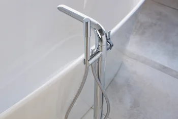 Chrome freestanding bath mixer with a handheld shower hose, mounted beside a white curved tub on light grey tiled flooring.