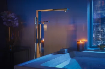 Modern chrome bathroom tap over a marble sink, bathed in blue light; candle by the window, purple flowers in a vase, city lights outside.