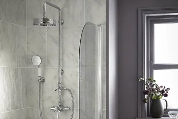 Modern bathroom shower with marble tile walls, chrome fixtures, and a curved glass screen; a window sill holds a dark vase with purple flowers.