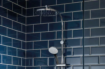 Blue tiled shower area with chrome rain shower overhead and a handheld attachment on a vertical rail; glass partition on the right.