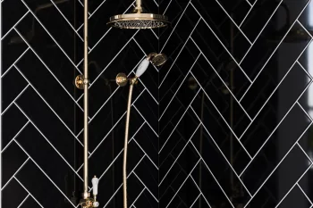 Gold-brass shower system with a large round rain head and handheld spray, mounted on a glossy black wall with white herringbone tiles.