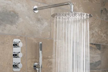 Chrome rainfall shower head with water cascading from a curved arm above beige tiled shower wall; handheld wand and chrome control panel sit beside.