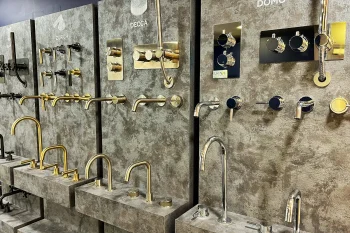 Showroom wall displaying gold and chrome bathroom taps, spouts and handles mounted on textured grey panels with brand labels.