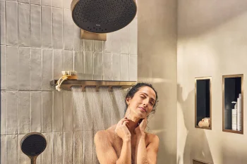 A person stands under a rainfall shower in a modern bathroom, eyes closed, hands at their neck; water flows from a large overhead shower and handheld unit.