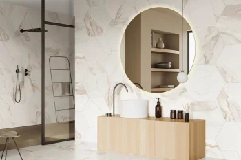 Marble-tiled bathroom with a round backlit mirror above a light-wood vanity, white vessel sink, dark faucet, and a black-framed glass shower to the left.